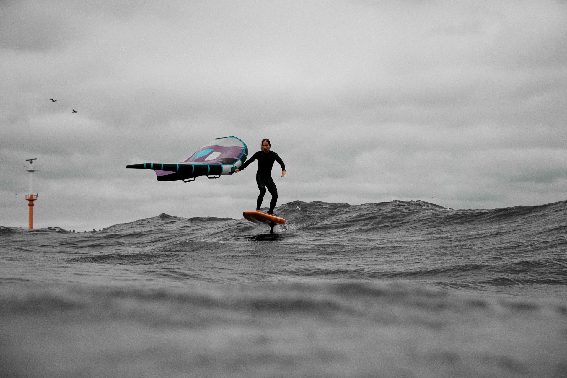 Surfeur en combinaison de plongée debout sur une planche au milieu des vagues, tenant les ailes pour le wingfoil d’une seule main